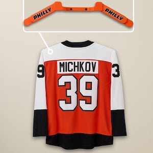 Pode incluir: Uma camisola de hóquei laranja e preta com o nome "MICHKOV" e o número "39" nas costas. A camisola tem mangas brancas com os números "9" e "3". Acima da camisola, um cabide laranja com a palavra "PHILLY".