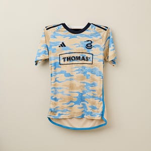 May include: A short-sleeved jersey with a camouflage pattern in tan and light blue. The jersey has a navy blue collar and trim, with the word "THOMAS" in a rectangular box on the front. The number "2" is visible on the upper right.
