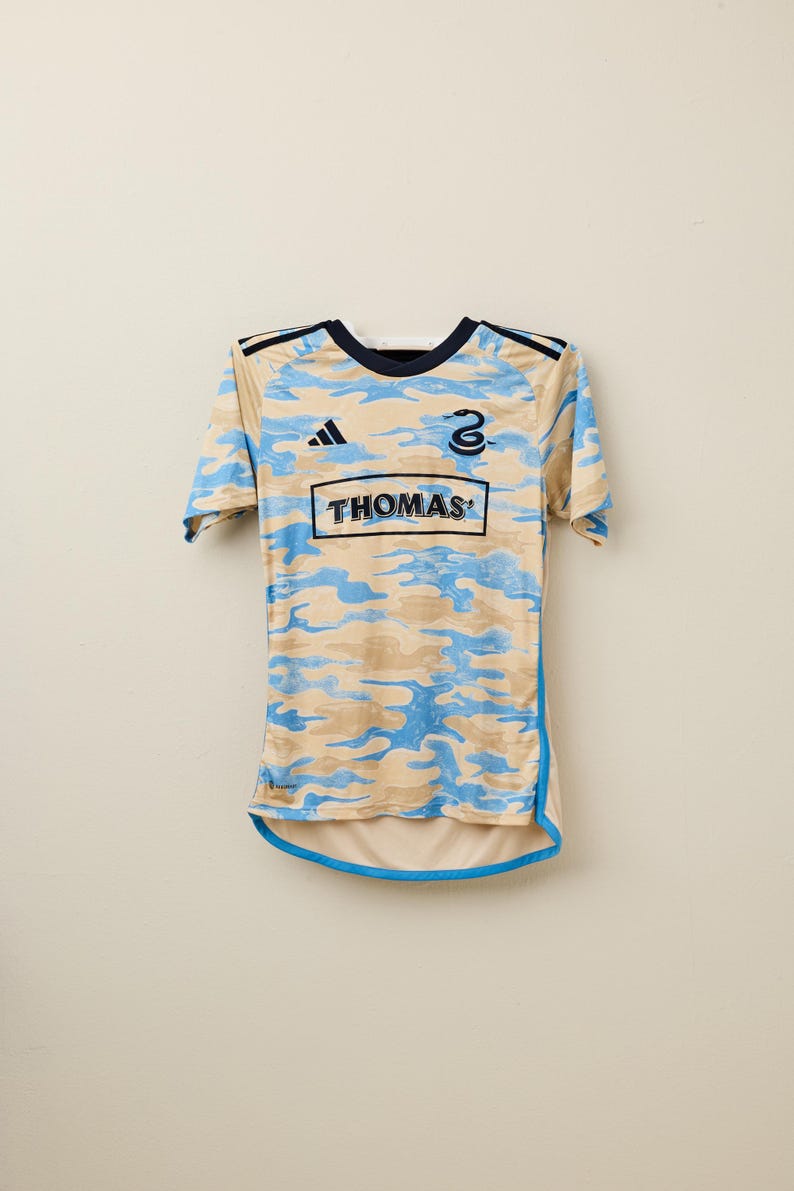 May include: A short-sleeved jersey with a camouflage pattern in tan and light blue. The jersey has a navy blue collar and trim, with the word "THOMAS" in a rectangular box on the front. The number "2" is visible on the upper right.