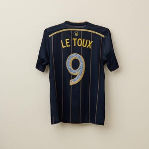 May include: Navy blue soccer jersey with gold vertical stripes. The back features the name "LE TOUX" in gold lettering above a large number "9" with a blue and gold design.