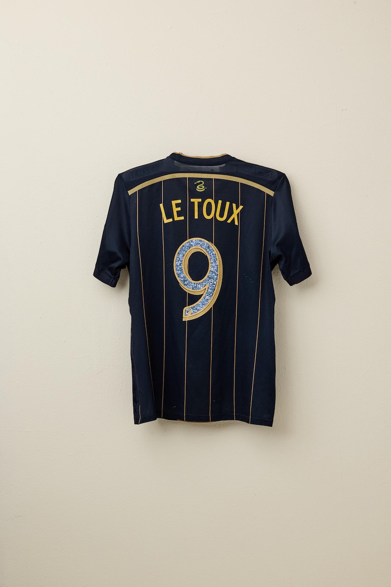 May include: Navy blue soccer jersey with gold vertical stripes. The back features the name "LE TOUX" in gold lettering above a large number "9" with a blue and gold design.