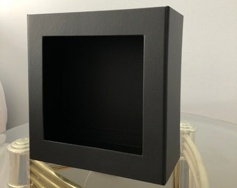 May include: A black square shadow box with a square opening in the front. The shadow box is sitting on a glass table with gold metal legs.