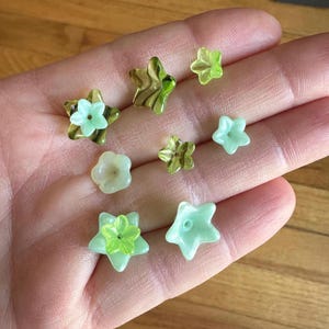 May include: A collection of eight green and white glass flower-shaped beads. The beads are in the shape of stars and flowers. The beads are on a hand.