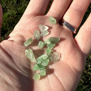 May include: A handful of small, translucent, light green crystals. The crystals vary in size and shape, with a slightly rough texture. The crystals are held in an open palm, with a silver ring visible on a finger. The background is blurred.