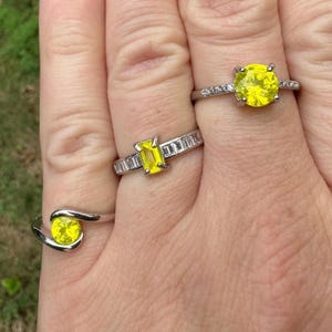 May include: Three silver rings with bright yellow gemstones. One ring features a round stone, another has a rectangular stone, and the third has a spiral design with a round stone. The rings are set against a blurred green background.