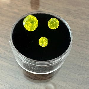 May include: Three faceted, bright yellow gemstones of varying sizes are displayed in a clear, round container with a black velvet interior. The gemstones are arranged in a triangular pattern, showcasing their brilliance.