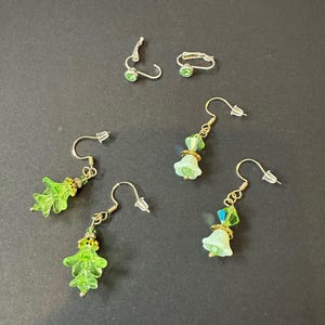 May include: A selection of handmade earrings with gold-coloured hooks and green glass beads. Some earrings have leaf-shaped pendants, others have bell-shaped or round green beads. There are also a pair of stud earrings and a small hoop earring.
