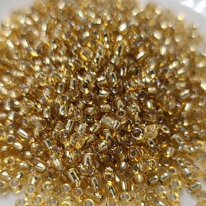 May include: A pile of small, gold-colored glass seed beads. The beads are round and have a metallic sheen.