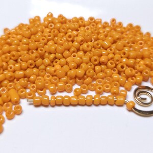 May include: A large pile of small, round, orange beads. The beads are scattered on a white surface. A small gold metal spiral is also visible in the image.