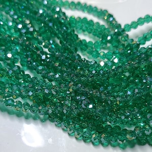 May include: Close-up of strands of emerald green faceted beads. The beads are small, round, and have a sparkling, iridescent quality. The beads are strung together, creating a flowing, textured appearance. The background is white.