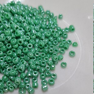 May include: A pile of small, round, green glass beads. The beads are shiny and have a slight iridescent sheen.
