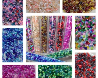 May include: Assortment of seed beads in various colors and finishes, including clear, iridescent, and opaque. The beads are displayed in small containers and loose piles.