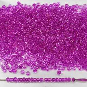 May include: A large quantity of small, clear, purple glass beads. The beads are round and have a slight sheen. A single strand of the beads is shown on a white surface.