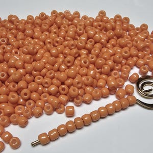 May include: A large pile of small, round, orange beads. The beads are scattered on a white surface. A gold metal beading needle is lying on top of the beads.