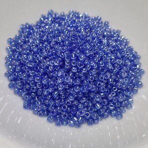 May include: A large pile of small, iridescent blue glass seed beads. The beads are round and have a smooth, shiny finish.