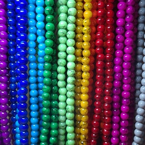 May include: A close-up of a rainbow assortment of colorful glass beads. The beads are strung together in long strands and are arranged in a vertical line. The beads are all the same size and shape, and they are all glossy and smooth.