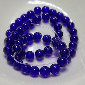 May include: A strand of deep blue glass beads. The beads are round and translucent, with subtle swirling patterns inside. The beads are strung on a clear, thin cord, and are arranged in a circular pattern.