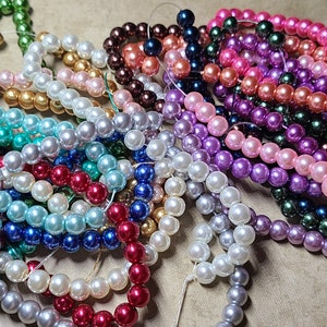 May include: A collection of beaded strands in various colors, including green, black, white, blue, red, and purple. The beads appear to be round and have a glossy finish. The strands are arranged in a pile, suggesting they are for jewelry making or crafts.
