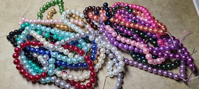 May include: A collection of beaded strands in various colors, including green, black, white, blue, red, and purple. The beads appear to be round and have a glossy finish. The strands are arranged in a pile, suggesting they are for jewelry making or crafts.