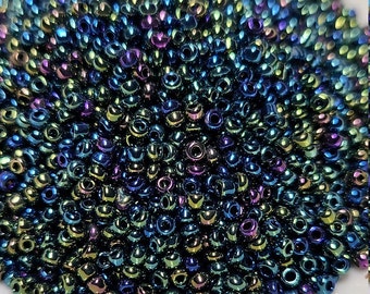 May include: A close-up shot of a pile of small, iridescent seed beads. The beads have a dark base color with shimmering hues of blue, green, and purple. They are densely packed together, creating a textured surface.