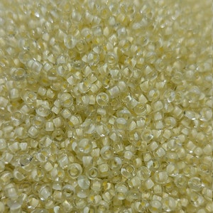 May include: A close-up image of a pile of small, translucent, round seed beads. The beads are a pale yellow color and have a slight iridescent sheen.