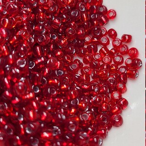 May include: A close-up shot of a pile of small, translucent red beads. The beads are densely packed together, reflecting light and creating a shimmering effect. The beads are likely used for jewelry making or other craft projects.