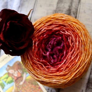 Peut inclure: Une pelote de laine avec un d&eacute;grad&eacute; de couleurs orange, rouge et rose. La laine est enroul&eacute;e en forme de boule et repose sur une surface en bois. Il y a une rose s&eacute;ch&eacute;e et une carte avec une image d'une femme sur la surface.