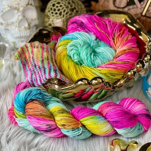 Puede incluir: Un ovillo de hilo de colores en tonos de rosa, azul, verde y amarillo. El hilo está enrollado alrededor de un cuenco dorado en forma de mano.