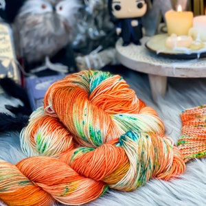 May include: A skein of orange, white, and green yarn with a small knitted swatch of the same yarn. The yarn is on a white fluffy surface.
