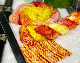 May include: A skein of yarn in shades of red, orange, and yellow, with a small knitted swatch in the same colors. The yarn is labeled "Dragon Fire".