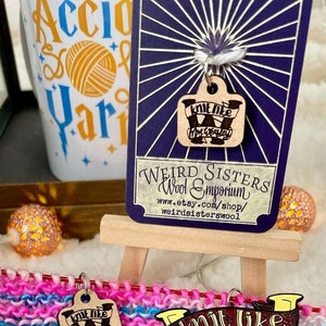 May include: Knitting stitch markers with a wooden tag that says "Knit like Mrs. Weasley". The markers are on a colorful knitted fabric.