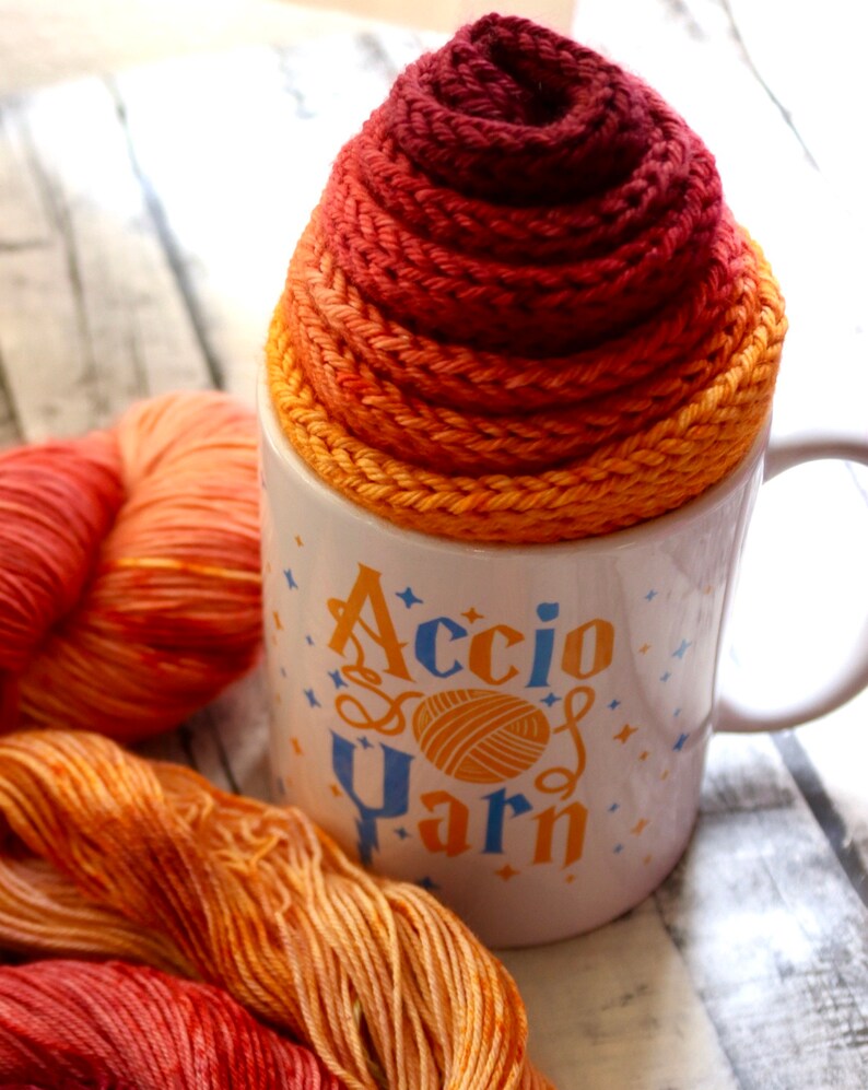 Peut inclure: Une tasse en c&eacute;ramique blanche avec le texte "Accio Yarn" en lettres bleues et orange. La tasse est surmont&eacute;e d'une pelote de laine orange, rouge et jaune.