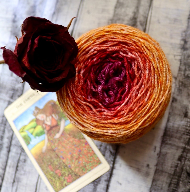 Peut inclure: Une pelote de laine avec un d&eacute;grad&eacute; de couleurs orange, rouge et rose. La laine est enroul&eacute;e en forme de boule et repose sur une surface en bois. Il y a une rose s&eacute;ch&eacute;e et une carte avec une image d'une femme sur la surface.