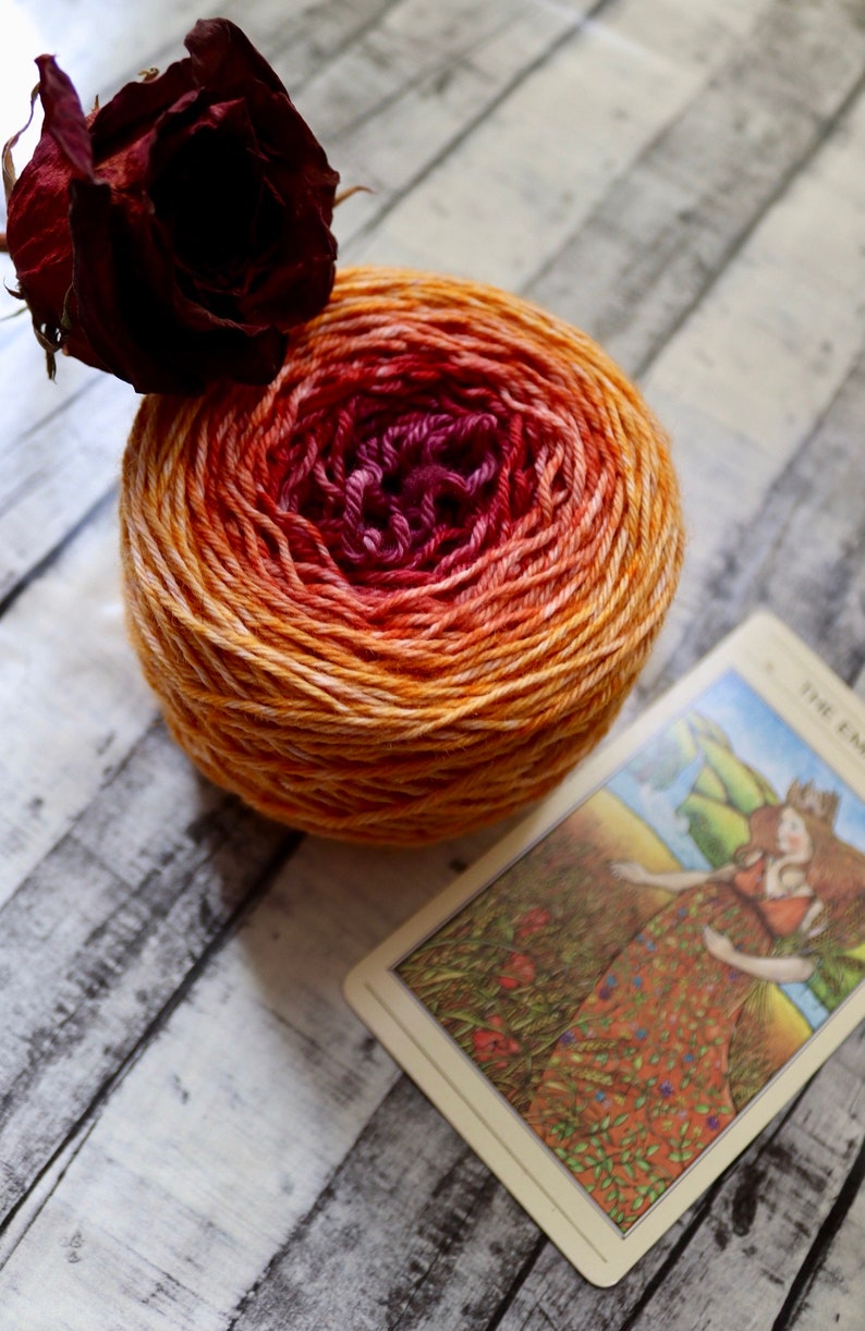 Peut inclure: Une pelote de laine avec un d&eacute;grad&eacute; de couleurs orange, jaune et rouge. Une rose s&eacute;ch&eacute;e est en arri&egrave;re-plan. Le texte "THE EMPRESS" est visible sur une carte au premier plan.