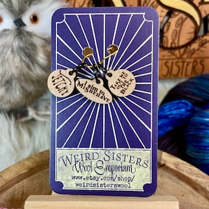 May include: A purple card with the text "Weird Sisters Wool Emporium" and a website address. Three wooden stitch markers with the text "Take me out to the black", "I aim to misbehave", and "Jetchy" are attached to the card.