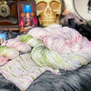 Pode incluir: Dois novelos de fio tingido &agrave; m&atilde;o em uma combina&ccedil;&atilde;o de cores branca, verde e rosa. O fio est&aacute; disposto em uma pilha sobre uma superf&iacute;cie cinza e felpuda. Um pequeno peda&ccedil;o de tecido de malha &eacute; vis&iacute;vel em primeiro plano.