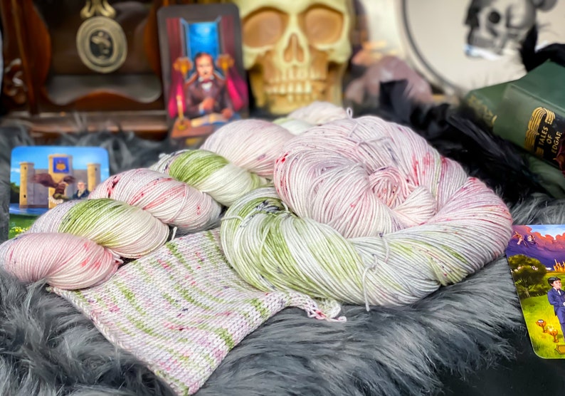 Pode incluir: Dois novelos de fio tingido &agrave; m&atilde;o em uma combina&ccedil;&atilde;o de cores branca, verde e rosa. O fio est&aacute; disposto em uma pilha sobre uma superf&iacute;cie cinza e felpuda. Um pequeno peda&ccedil;o de tecido de malha &eacute; vis&iacute;vel em primeiro plano.