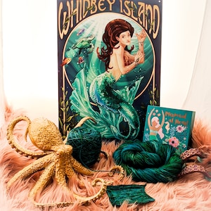 May include: A gold octopus figurine with a green yarn ball and a skein of green yarn. The octopus is on a pink fuzzy surface. A card with the text "Mermaid at Heart" is in the background.