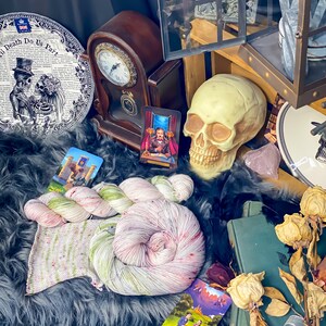 Pode incluir: Uma natureza morta com uma variedade de itens tem&aacute;ticos g&oacute;ticos e assustadores, incluindo um rel&oacute;gio, uma caveira, um prato com um design de caveira e ossos cruzados, cartas de tarot e um novelo de fio. Os itens est&atilde;o dispostos em uma superf&iacute;cie de pele preta.