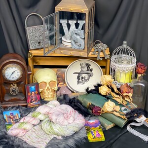Pode incluir: Uma natureza-morta com uma variedade de itens vintage e decorativos, incluindo uma caixa de madeira, um rel&oacute;gio, uma caveira, uma gaiola, um prato com um desenho de caveira, um livro e l&atilde;. Os itens est&atilde;o dispostos sobre uma superf&iacute;cie preta com um fundo branco.