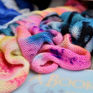 Puede incluir: Primer plano de hilo y tela de punto hechos a mano y coloridos. El hilo y la tela están teñidos en un arcoíris de colores, incluyendo rosa, amarillo, azul y morado. La tela de punto es suave y texturizada, con una variedad de colores y patrones.