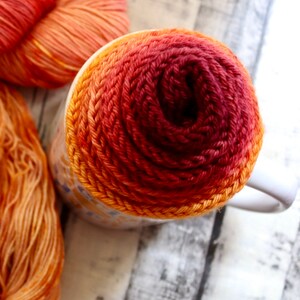 Peut inclure: Gros plan d'une tasse envelopp&eacute;e de fil orange, rouge et bordeaux. Le fil est enroul&eacute; en spirale, cr&eacute;ant un effet d&eacute;grad&eacute;.