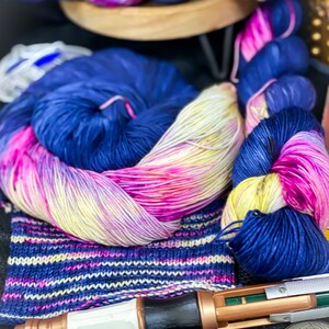 May include: A close-up of colorful yarn in shades of pink, blue, and yellow. The yarn is arranged in a pile and a skein. A knitted sock is visible in the foreground, along with a small, brown and black object.