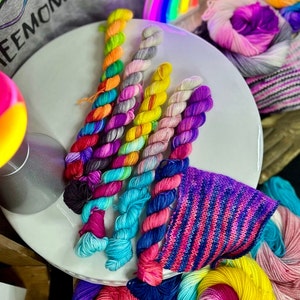 May include: A close-up of a collection of colorful yarn skeins, featuring a variety of bright colors and patterns. The yarn is arranged in a circular pattern on a white surface.