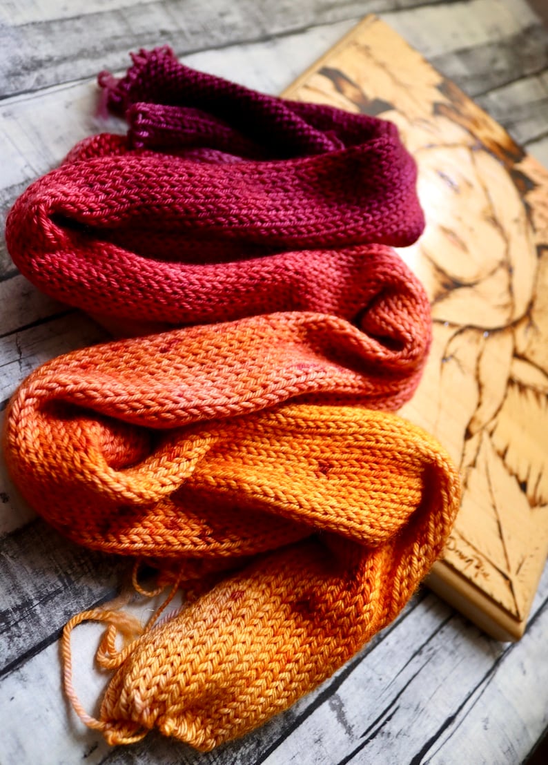 Peut inclure: Trois &eacute;charpes en tricot dans des tons d'orange, de rouge et de violet. Les &eacute;charpes sont empil&eacute;es les unes sur les autres, l'&eacute;charpe orange en bas, l'&eacute;charpe rouge au milieu et l'&eacute;charpe violette en haut.