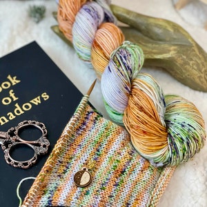 Op de afbeelding: Een close-up van een bol garen met een veelkleurig patroon van wit, oranje, groen en paars. Het garen is om een houten hand gewikkeld en op de voorgrond ligt een breiproject. Het breiproject is een gestreept patroon van wit, oranje, groen en paars. Een kleine houten maanbedel is aan het breiproject bevestigd.