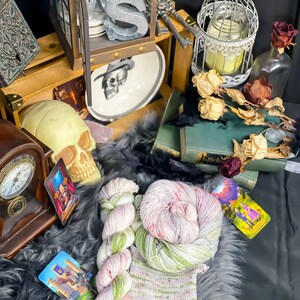 Pode incluir: Dois novelos de fio tingido &agrave; m&atilde;o em tons de verde, rosa e branco. O fio est&aacute; disposto sobre uma superf&iacute;cie cinza e felpuda com uma pequena amostra de tric&ocirc; no centro. H&aacute; cartas de tarot e outros itens decorativos no fundo.
