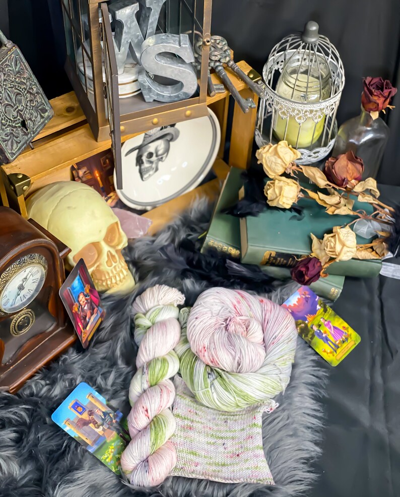 Pode incluir: Dois novelos de fio tingido &agrave; m&atilde;o em tons de verde, rosa e branco. O fio est&aacute; disposto sobre uma superf&iacute;cie cinza e felpuda com uma pequena amostra de tric&ocirc; no centro. H&aacute; cartas de tarot e outros itens decorativos no fundo.