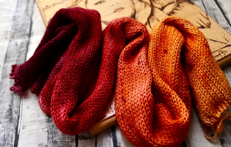 Peut inclure: Trois &eacute;charpes en tricot dans des tons de rouge, orange et jaune. Les &eacute;charpes sont faites avec un point textur&eacute; et sont dispos&eacute;es en pile sur une surface en bois.