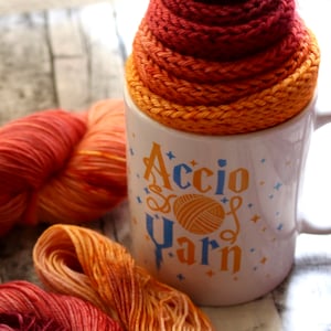 Peut inclure: Une tasse en c&eacute;ramique blanche avec le texte "Accio Yarn" en lettres bleues et jaunes. La tasse est remplie d'une pelote de laine dans des tons d'orange, de jaune et de rouge.